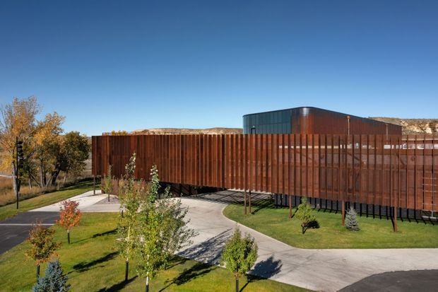 Exterior view of a contemporary low-rise building set within landscaped grounds. A long elevated wing is enclosed by vertical rust-coloured metal cladding and screened façades, with a darker glazed and metal upper volume behind it. Curving paved paths, lawn, young trees, and autumn vegetation surround the structure under a clear blue sky.