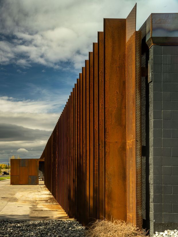 Exterior architectural photograph of a long, dramatic facade composed of repeated vertical weathered steel fins and large rusted steel panels. A dark grey concrete-block wall and perforated metal screen form part of the building edge. The linear facade recedes strongly into the distance beside a paved walkway, with a cloudy sky and sparse landscape in the background. The composition emphasizes industrial materials, repetition, texture, and angular geometry.