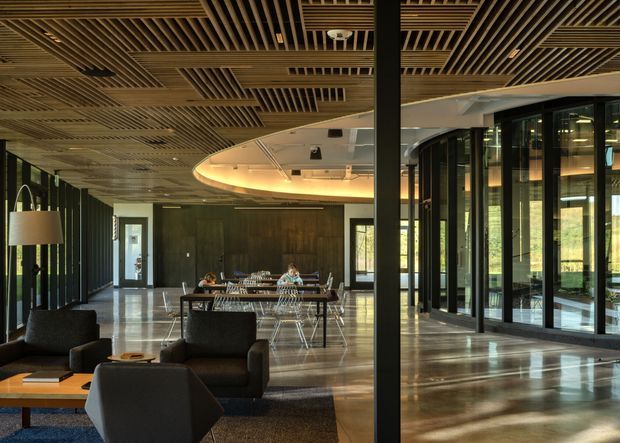 A spacious contemporary commercial interior with a lounge area in the foreground and a communal dining or meeting area beyond. The space features dark-framed floor-to-ceiling glazing, polished concrete flooring, a dramatic curved suspended ceiling feature, exposed services, and an extensive slatted timber ceiling creating a warm textural contrast with the dark metal structure.