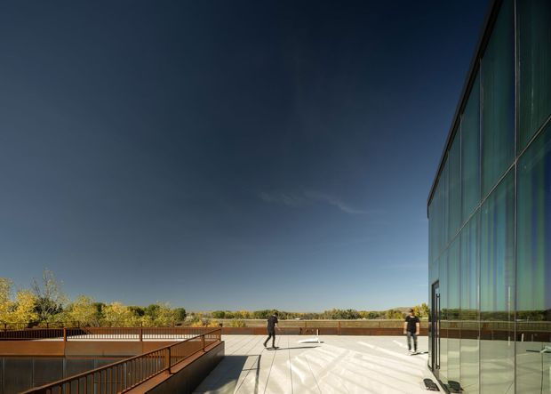 A broad rooftop or elevated terrace beside a tall glazed building facade. The space has pale paved flooring, dark metal balustrades, and a long open view toward a low tree-lined horizon. Two people provide scale, while the reflective curtain-wall glazing dominates the right side of the image. The scene has a contemporary institutional or commercial character and strong late-day light.