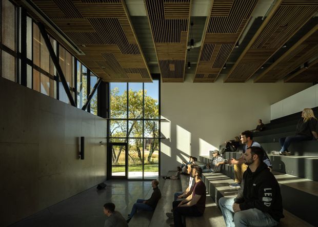 A modern multi-purpose lecture or presentation space with tiered stepped seating occupied by people, a large glazed doorway and windows opening to trees outside, and a dramatic timber slatted acoustic ceiling. The room has dark polished flooring, a tall concrete or grey wall, exposed structural framing, and strong natural sunlight.