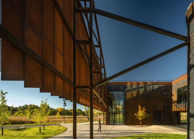 Exterior architectural view of a contemporary low-rise building with a dramatic rusted steel mesh and plate facade, exposed structural steel bracing, and a glazed entrance volume. The building forms a sheltered paved courtyard or passage beside landscaped lawn and young trees, with strong late-day sunlight and clear blue sky.