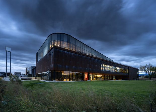 Exterior dusk photograph of a large contemporary commercial building with a dark metal-clad lower volume, extensive illuminated curtain-wall glazing, and a distinctive curved, sloping glazed upper roof or clerestory form. The building sits within a broad landscaped lawn under a dramatic cloudy sky.