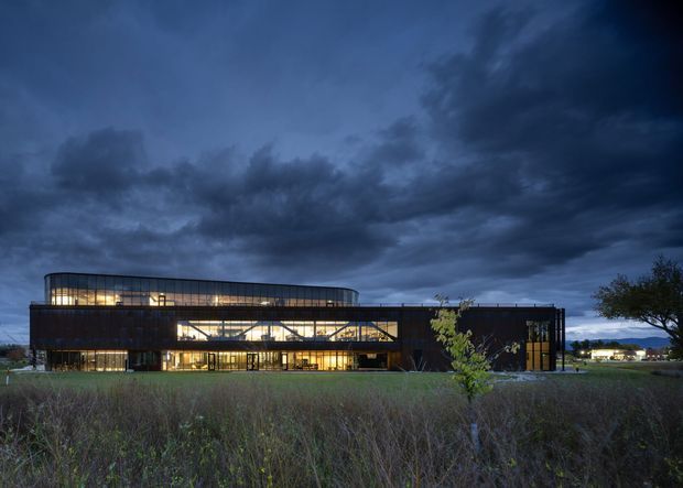 A large contemporary institutional or commercial building photographed at dusk, set within an open landscaped field. The building has a long, low form with dark cladding, extensive illuminated floor-to-ceiling glazing, and a gently curved upper roofline. Dramatic blue-grey clouds dominate the sky, while warm interior lighting creates a strong contrast against the dark exterior.