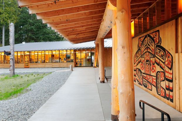 Covered timber walkway beside a low-rise building, featuring massive round wood posts, exposed timber roof framing, vertical timber wall cladding, and a large Indigenous-style graphic mural. Glazed rooms with warm interior lighting are visible across the courtyard, while gravel landscaping and carved timber posts frame the exterior setting.