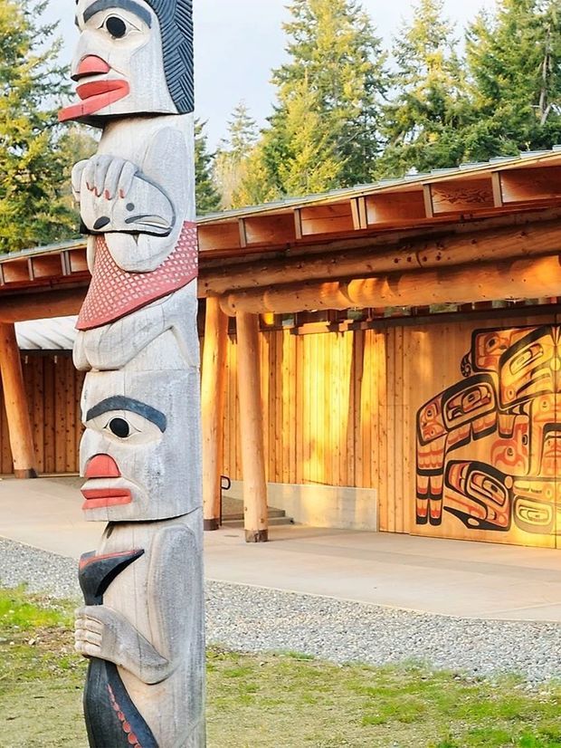 Outdoor view of a timber-clad cultural or community structure with a low pitched roof, exposed round-log posts and beams, and prominent Indigenous carved and painted artwork. A tall carved totem pole stands in the foreground beside a covered concrete-paved area. The setting is surrounded by trees and gravel landscaping.