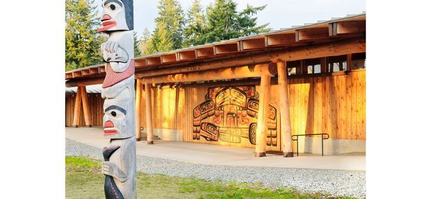 Exterior view of a low, timber-clad pavilion or cultural building beside a carved and painted totem pole. The structure features warm natural wood siding, exposed timber posts and beams, a shallow sloping roof with broad eaves, and a large colourful Indigenous-inspired mural or carving on the rear wall. Gravel surrounds the building, with trees in the background.