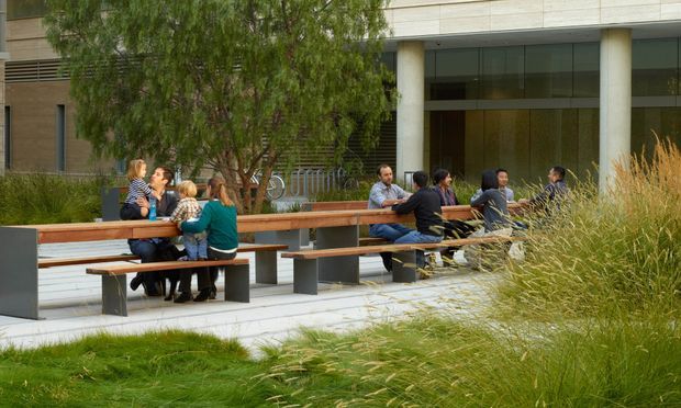 A landscaped outdoor courtyard or plaza beside a contemporary institutional or commercial building. Long timber-topped communal tables with dark metal supports provide seating for groups, surrounded by ornamental grasses and mature planting. The building behind has a restrained modern facade with large glazed openings, pale vertical columns, horizontal cladding, and shaded recessed areas.