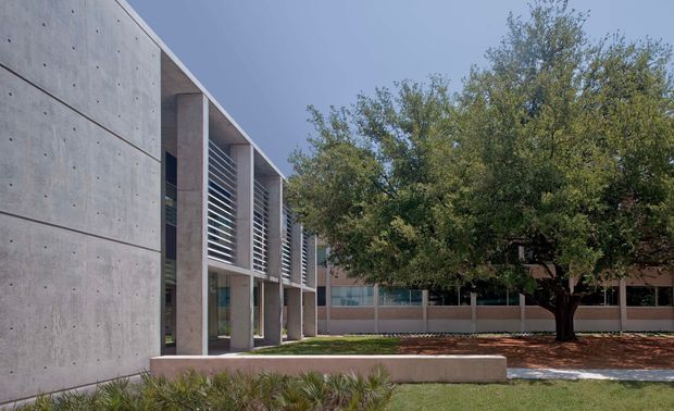 Exterior architectural view of a contemporary institutional or commercial building. The composition shows a long exposed concrete facade with vertical structural bays, recessed glazing, and horizontal metal louvres. A landscaped courtyard with mature trees and low concrete retaining walls sits alongside the building.