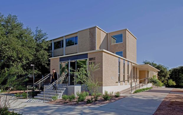 Exterior view of a contemporary multi-storey brick building with extensive glazing, projecting upper-level volumes, a perforated brick screen, accessible ramp and stairs, and landscaped surroundings. The building appears institutional or commercial in character.