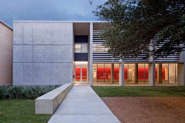 Contemporary two-storey building exterior viewed from a landscaped front approach. The facade combines large smooth concrete panels, slender concrete columns, extensive glazed doors and windows, and horizontal louvres or screens across the upper level. Warm interior lighting and vivid red blinds or panels are visible through the glazing. A minimalist concrete path and low concrete retaining bench lead across a lawn toward the entrance.
