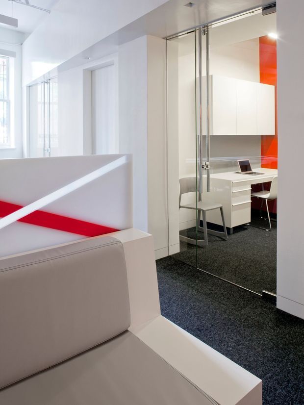 Bright contemporary office interior with a white and red colour scheme. The foreground contains built-in white seating with a red inset feature, while a glass sliding door reveals a compact private workstation with white cabinetry, desk drawers, chair and laptop. Dark carpet contrasts with the white walls and joinery, and large windows provide abundant natural light.