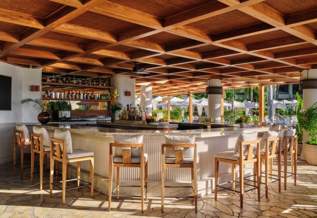 A bright, open-air hospitality bar with a long curved counter, high stools, and extensive bottle shelving. The space features a warm timber coffered ceiling with woven infill panels, timber bar stools, a ribbed light-coloured bar front, stone-patterned flooring, and large open glazed areas connecting to an outdoor setting.