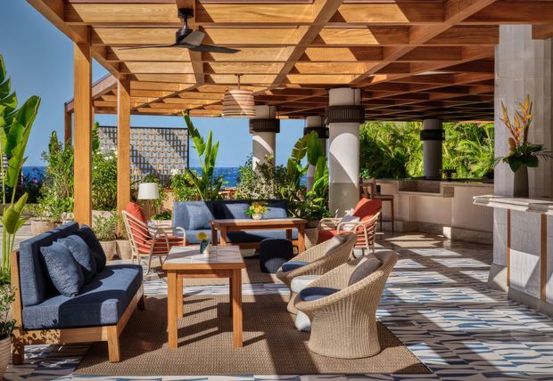 A luxurious outdoor covered entertaining area with a timber pergola and exposed timber ceiling structure, supported by substantial columns. The space includes multiple lounge seating groups, timber coffee tables, woven armchairs, patterned outdoor flooring, lush tropical planting, pendant lighting, and a built-in outdoor kitchen or bar along the rear wall. Ocean and distant landscape are visible beyond the open sides.