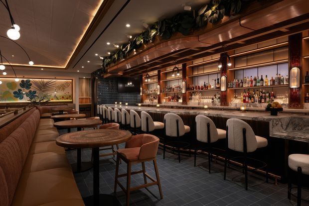 A sophisticated restaurant or cocktail bar interior with a long marble-topped bar, extensive backlit bottle shelving, upholstered bar stools, and a built-in banquette with round timber tables. The space features dark tiled flooring, warm timber and bronze-toned joinery, layered pendant and recessed lighting, decorative greenery above the bar, and a large botanical wall artwork. The atmosphere is intimate, warm, and refined.