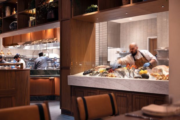 A warmly lit upscale commercial restaurant interior featuring an open kitchen and chef's counter. Dark timber cabinetry and wall panelling frame a large pale stone seafood display counter, with chefs working behind it. White rectangular tiled walls, stainless-steel kitchen equipment, recessed lighting, and leather dining chairs are visible.