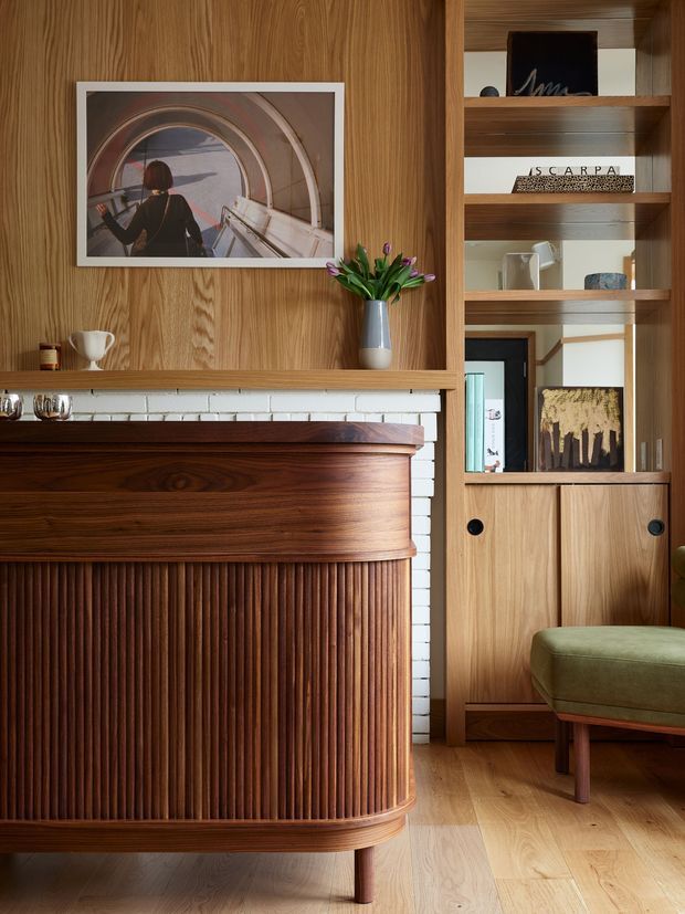 A warmly styled living room interior featuring a richly grained timber wall, built-in timber shelving and cabinetry, and a sculptural fluted timber sideboard or bar cabinet. A white painted brick backsplash is visible behind the cabinet, with framed artwork, books, ceramics, and a green upholstered occasional chair adding character. Light timber flooring and warm natural light create a refined mid-century-inspired atmosphere.