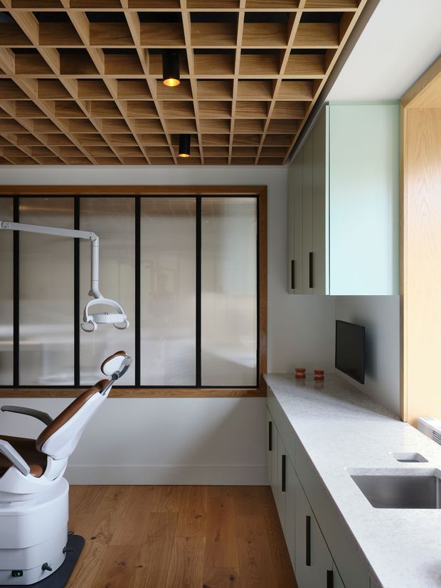 Bright, compact dental treatment room with a dental chair and overhead examination light. The room has pale cabinetry with dark handles, a light stone-look benchtop and integrated sink, a wall-mounted monitor, frosted black-framed glazing, and a distinctive timber coffered ceiling with recessed downlights. Warm timber flooring and restrained finishes create a clean, contemporary clinical interior.