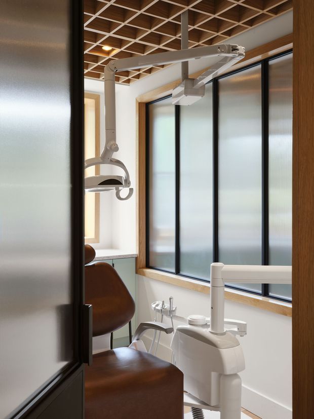 A compact, contemporary dental treatment room viewed through a doorway, featuring a brown dental chair and white dental delivery unit beneath articulated examination lights. Full-height frosted glazing with dark frames and timber surrounds lines one wall, while the ceiling has a distinctive exposed timber grid with white cross members. The space is bright, clean, and carefully detailed.