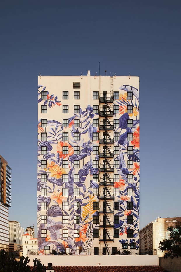 Exterior view of a tall urban apartment or commercial building with a cream-painted facade covered in a large-scale colorful botanical mural. The mural features oversized purple, blue, orange, red, and yellow leaves and flowers around rows of black-framed windows. A prominent black metal external fire-escape stair runs vertically down the center-right of the facade. The building is photographed against a clear dark blue sky, with surrounding city buildings partially visible at the edges and a low rooftop wall in the foreground.