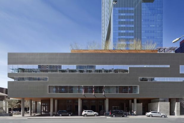 Street-level exterior view of a large contemporary multi-storey commercial building. The lower podium has a dark grey grid-like cladding system, extensive horizontal bands of reflective glazing, a recessed ground-level colonnade and entry zone, while a taller blue-glazed tower rises behind it. Vehicles and pedestrians are present along the street frontage.
