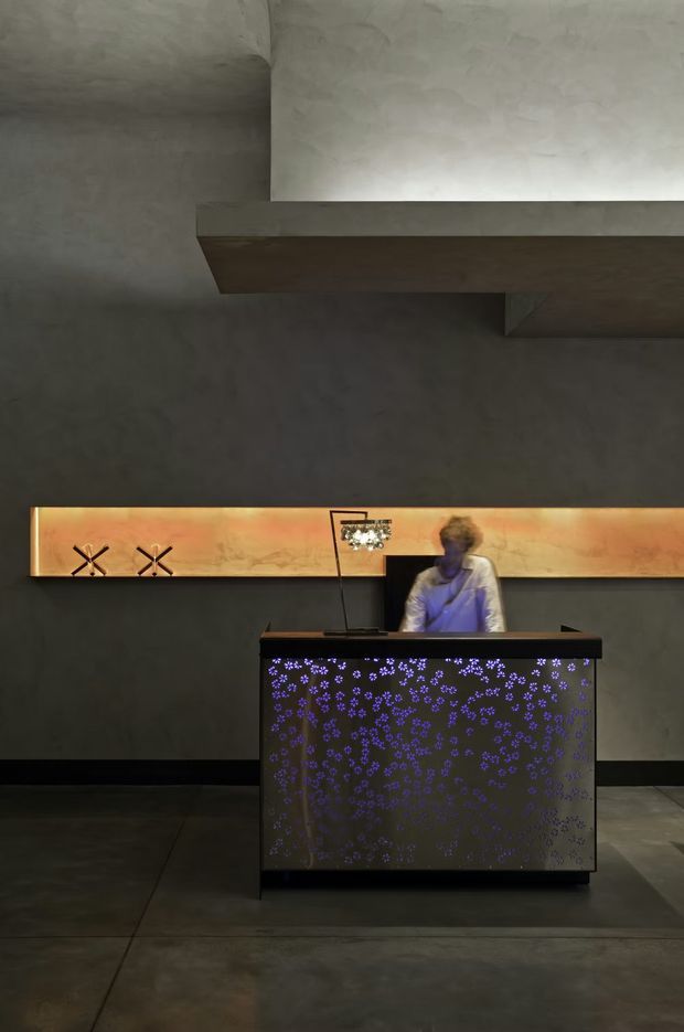 A dimly lit commercial reception or concierge area with a dark, industrial-style interior. A black reception desk with a perforated or patterned front panel illuminated by purple light stands in the foreground, with a staff member behind it. A long warm backlit timber or amber-toned feature panel spans the rear wall, while a large projecting ceiling bulkhead and dark concrete-like walls and floor create a dramatic architectural setting.