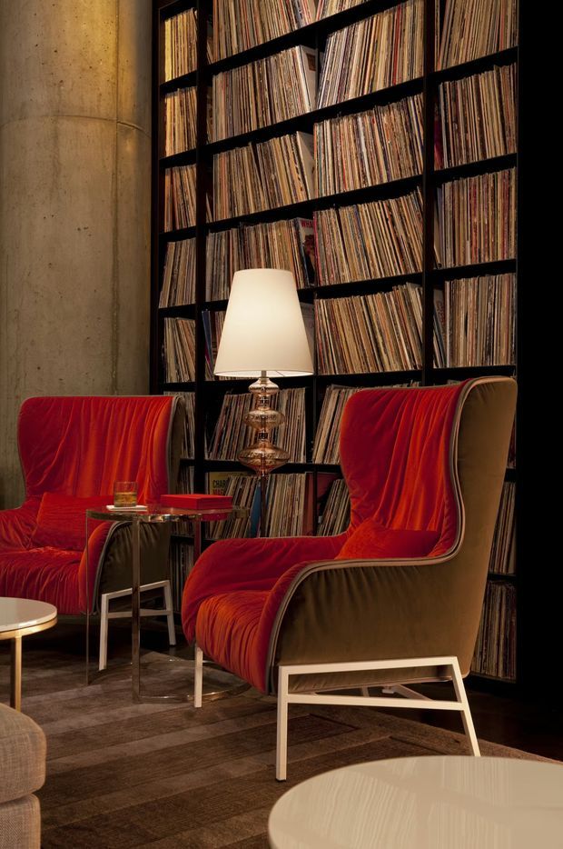 A moody, warmly lit music listening lounge with two sculptural red upholstered lounge chairs, a glass-and-metal side table, and a table lamp. A full-height black built-in shelving wall densely displays vinyl records, set against a raw cylindrical concrete column. Dark flooring and neutral furnishings create a refined, retro-inspired atmosphere.