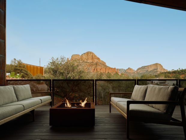 A covered outdoor terrace or balcony with dark timber decking, low black-framed glass balustrades, and a central square outdoor fire table. Two light upholstered lounge sofas face the fire, with a dramatic red-rock mountainous landscape visible beyond. The space has a warm, modern lodge-like atmosphere and strong indoor-outdoor connection.
