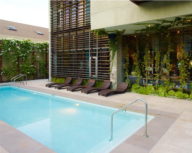 A landscaped courtyard pool area beside a contemporary building. A pale blue swimming pool with stainless-steel handrails occupies the foreground, bordered by light paving and dark timber loungers. The building features large glazed openings, a dense horizontal timber or metal screen, pale rendered wall panels, climbing greenery, and warm string lighting. A planted boundary screen and neighbouring roof are visible in the background.