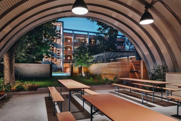 A covered communal outdoor dining and gathering area beneath a large ribbed arched canopy. Long timber-topped picnic tables with dark metal frames occupy the paved space, while timber screening, planted garden beds, trees, and a multi-storey building surround the courtyard. Pendant lights hang from the curved ceiling, creating a warm evening atmosphere.