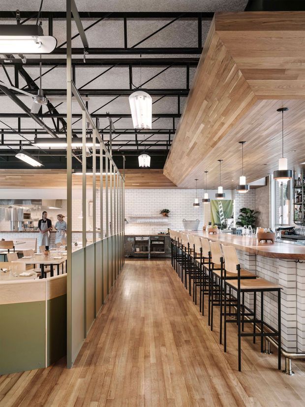 Interior view of a contemporary restaurant or café, featuring a long timber-topped bar with rows of stools, white brick-tile wall cladding, extensive timber wall and ceiling lining, exposed black structural framing, industrial pendant and track lighting, and a polished timber floor. A partially screened dining area and service counter are visible to the left.