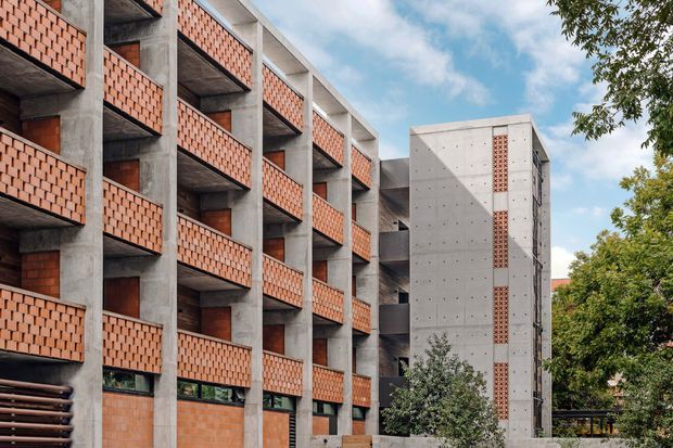 Exterior view of a multi-storey contemporary apartment building with exposed concrete structural frames and repeated balconies enclosed by terracotta brick screens. A contrasting concrete circulation or service tower features narrow vertical brick detailing. Mature trees and sky surround the urban residential complex.