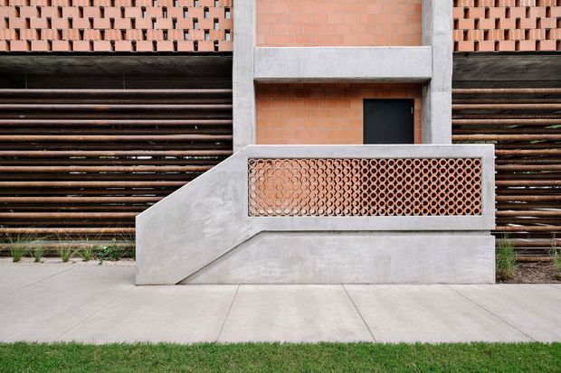 Exterior architectural detail featuring a terracotta brick facade with patterned perforated brickwork, dark horizontal timber or metal screening, exposed concrete structural beams, and a sculptural concrete stair or ramp enclosure with a perforated brick balustrade. A paved walkway and narrow lawn border the building.