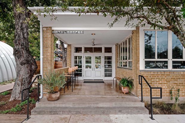 Front entrance of a low-rise brick building with a covered porch, broad concrete steps, black metal handrails, glazed double doors, large windows, and outdoor café-style seating. Mature trees and planted pots frame the entry, while a sign above the doors reads “HOTEL COFFEE.”