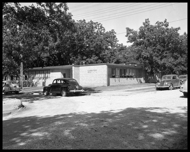 Black-and-white archival exterior photograph of a low, single-storey mid-century institutional or commercial building set among mature trees. The long horizontal composition features a flat roof, pale brick central entrance wall with small lettering, extensive horizontal window bands, and a shaded paved forecourt with several vintage cars.