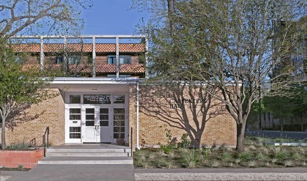 Exterior view of a brick civic or institutional building entrance, with a projecting upper level featuring patterned brickwork, slim metal or concrete columns, and a white glazed entry door beneath a shallow canopy. Steps, accessibility handrails, landscaped planting, and mature trees frame the facade. Signage identifies the building as a hall, though the full text is partially obscured.