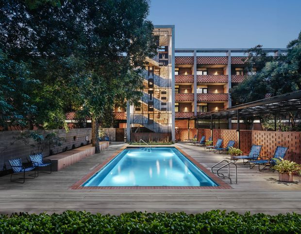 A landscaped courtyard pool area at dusk, framed by mature trees and surrounded by a multi-storey brick building. The rectangular swimming pool is set within a broad timber deck, with lounge chairs, built-in bench seating, brick privacy walls, and a lightweight covered outdoor structure. A glazed vertical stair or circulation tower and open balcony galleries are visible at the rear, creating a hotel or apartment-style courtyard setting.