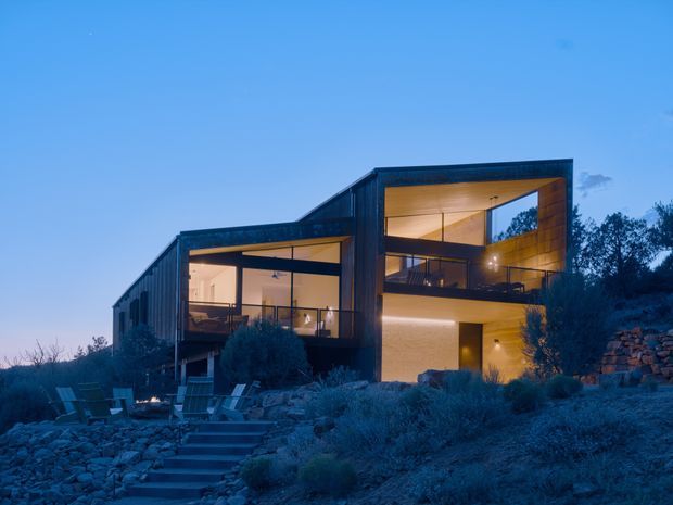 Contemporary multi-level house set into a rocky, landscaped hillside at dusk. The dark-clad rectangular structure features extensive floor-to-ceiling glazing, warm timber-lined interiors, projecting open terraces, and a prominent double-height glazed living area. Stone landscaping and steps lead toward outdoor seating in the foreground.