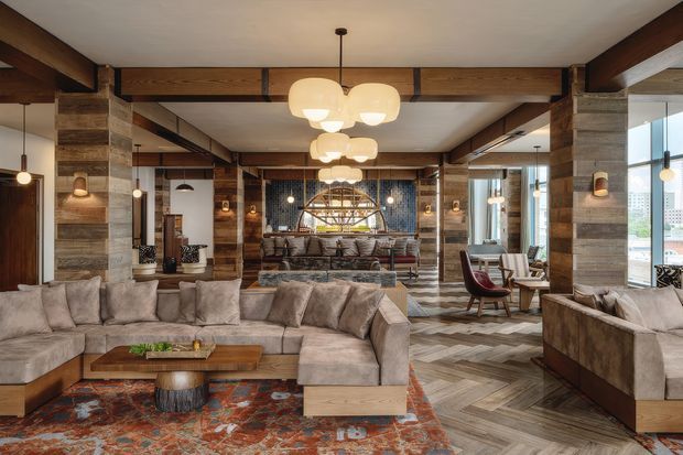 A spacious, richly textured open-plan lounge with a large sectional sofa, patterned rug, timber coffee table, lounge chairs, and layered pendant lighting. The room features substantial exposed timber beams and posts, reclaimed-looking timber wall cladding, dark mosaic tile detailing, chevron timber flooring, and expansive glazing. The overall design is warm, rustic, and lodge-inspired with a refined hospitality feel.