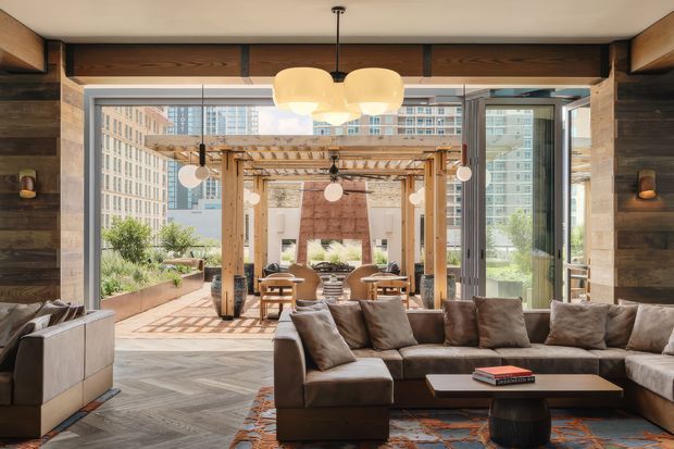 A warm contemporary living room with a large sectional sofa, low coffee table, and pendant lighting, opening through expansive glazed folding doors to a timber pergola and furnished outdoor terrace. Dark timber wall cladding, exposed timber framing, and a chevron-patterned timber floor create a rich, natural interior, with an urban high-rise setting visible beyond.