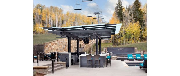 Outdoor hospitality and recreational terrace beneath a large slatted metal canopy, with bar-style counters, dining furniture, lounge seating, stone-clad walls, and ski-lift chairs visible overhead. The setting is surrounded by autumn forest and appears associated with a ski resort or mountain recreation facility.