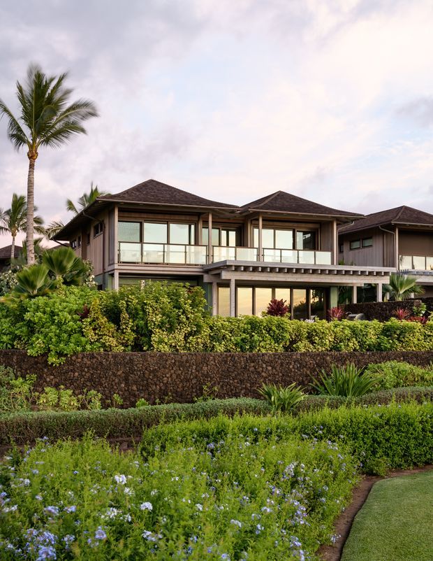 Exterior view of a large tropical-resort-style residence with multiple dark hipped roof forms, extensive glazing, covered outdoor terraces, and a landscaped foreground. The home is set behind layered hedges, flowering planting, palm trees, and a dark stone retaining wall.
