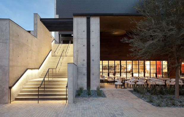 Exterior architectural view of a contemporary commercial building, featuring a broad illuminated concrete stairway, monumental concrete walls and columns, dark cladding, and a sheltered ground-floor restaurant or hospitality space with extensive black-framed glazing. A paved forecourt with built-in planting and outdoor tables occupies the foreground.