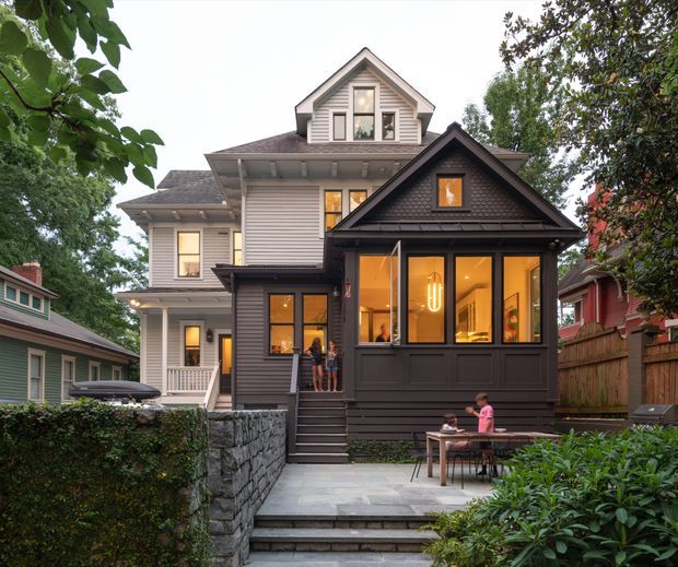 Exterior view of a multi-storey suburban residence with a prominent dark-clad gabled front extension or enclosed porch attached to a larger light-coloured weatherboard house. The facade features layered pitched roofs, horizontal siding, dark-framed windows, illuminated interiors, front steps, a paved terrace with outdoor dining, stone retaining walls, fencing, and established garden planting.