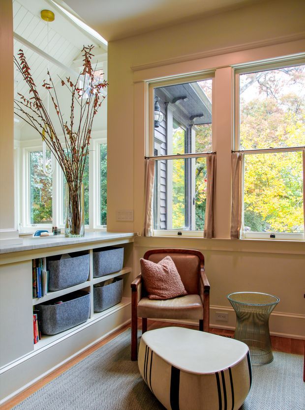 A bright, warm reading nook or compact living area with large double-hung windows, white built-in shelving and storage baskets, a pale countertop, and a leather armchair. A sculptural coffee table, metal side table, patterned carpet, and tall branch arrangement add contemporary decorative detail. The room has classic trim and plentiful natural light overlooking trees.