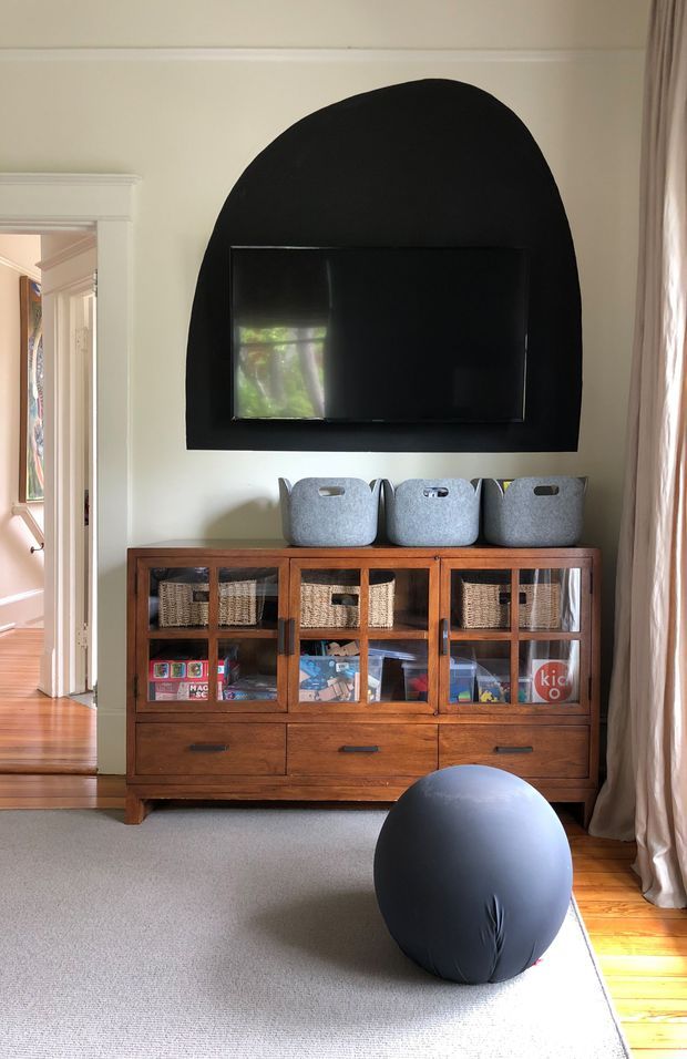 A warmly lit family living room with a wall-mounted television set within a bold black arched feature panel. Below it is a substantial mid-century-style timber storage cabinet with glass-fronted compartments, woven baskets, and children's items. Grey felt storage bins sit on top, while a large dark exercise ball rests on a pale carpet beside exposed timber floorboards. Cream walls, traditional trim, and full-length curtains complete the room.