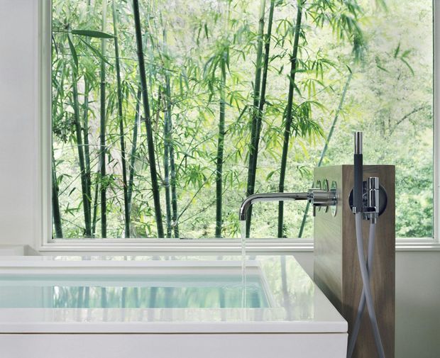 Close-up of a freestanding white bathtub positioned beside a large picture window overlooking dense bamboo and greenery. A minimalist wall-mounted or floor-mounted bath filler and handheld shower are set against a vertical dark timber panel. The bathroom has a bright, serene, nature-connected feel with clean contemporary lines.