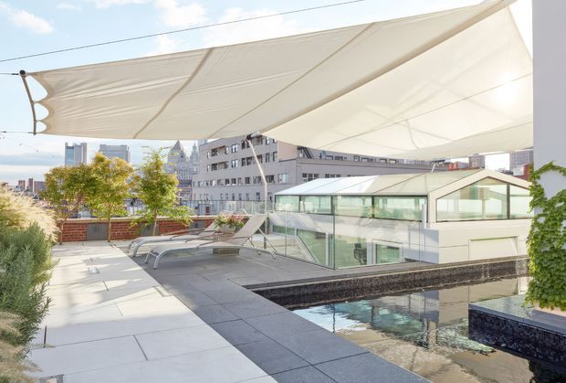 A bright rooftop terrace in an urban setting, featuring large white tensile shade sails, pale paved flooring, lounge chairs, landscaped planters, and a reflective rectangular water feature or pool. A contemporary glazed pavilion and surrounding city buildings are visible beyond the terrace.