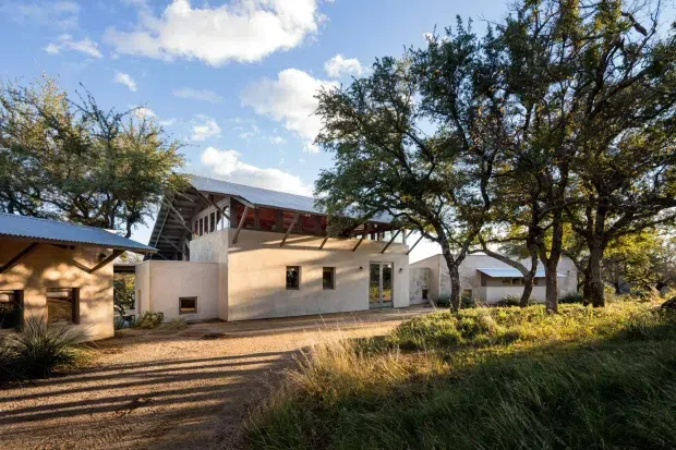 Exterior view of a contemporary low-rise residence or retreat set within a mature, dry landscaped setting. The building has pale rendered walls, broad metal pitched roofs, exposed timber roof framing and posts, small rectangular windows, and a glazed door opening onto a paved forecourt. Several connected or adjacent structures are visible among established trees and grasses.