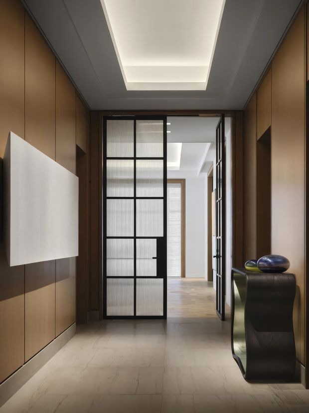 A refined interior entry corridor with warm wood wall panelling, pale stone flooring, and a recessed illuminated ceiling feature. A black-framed glass partition door opens toward a brighter adjoining space with additional wood flooring and built-in cabinetry. A sculptural dark console with decorative objects sits along the right wall, while a large minimalist white wall panel hangs on the left.